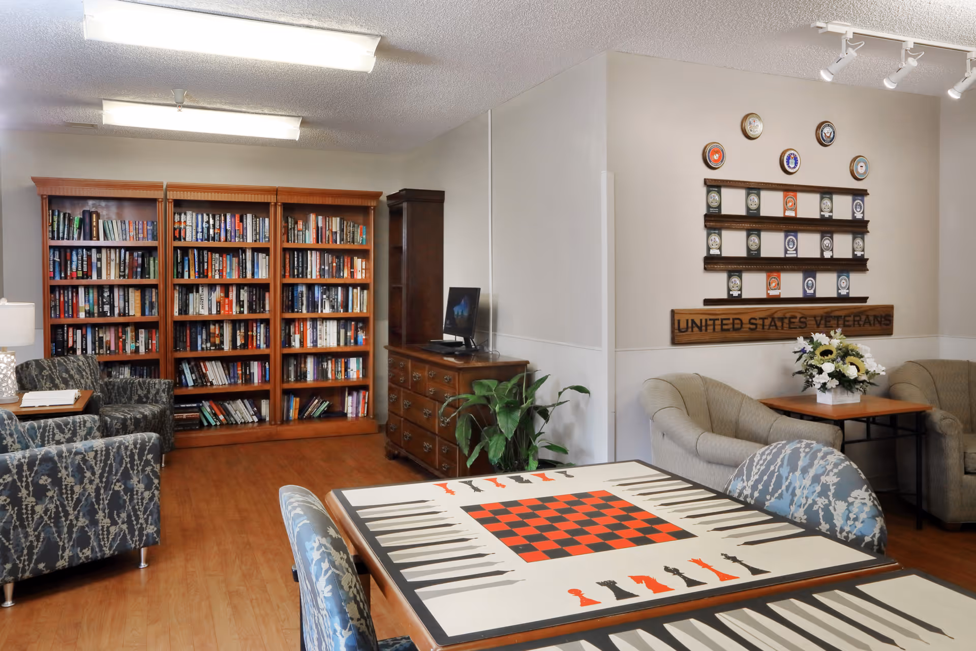 A cozy common room featuring bookshelves, comfortable chairs, a table with a checkerboard design, and a 'United States Veterans' wall display.