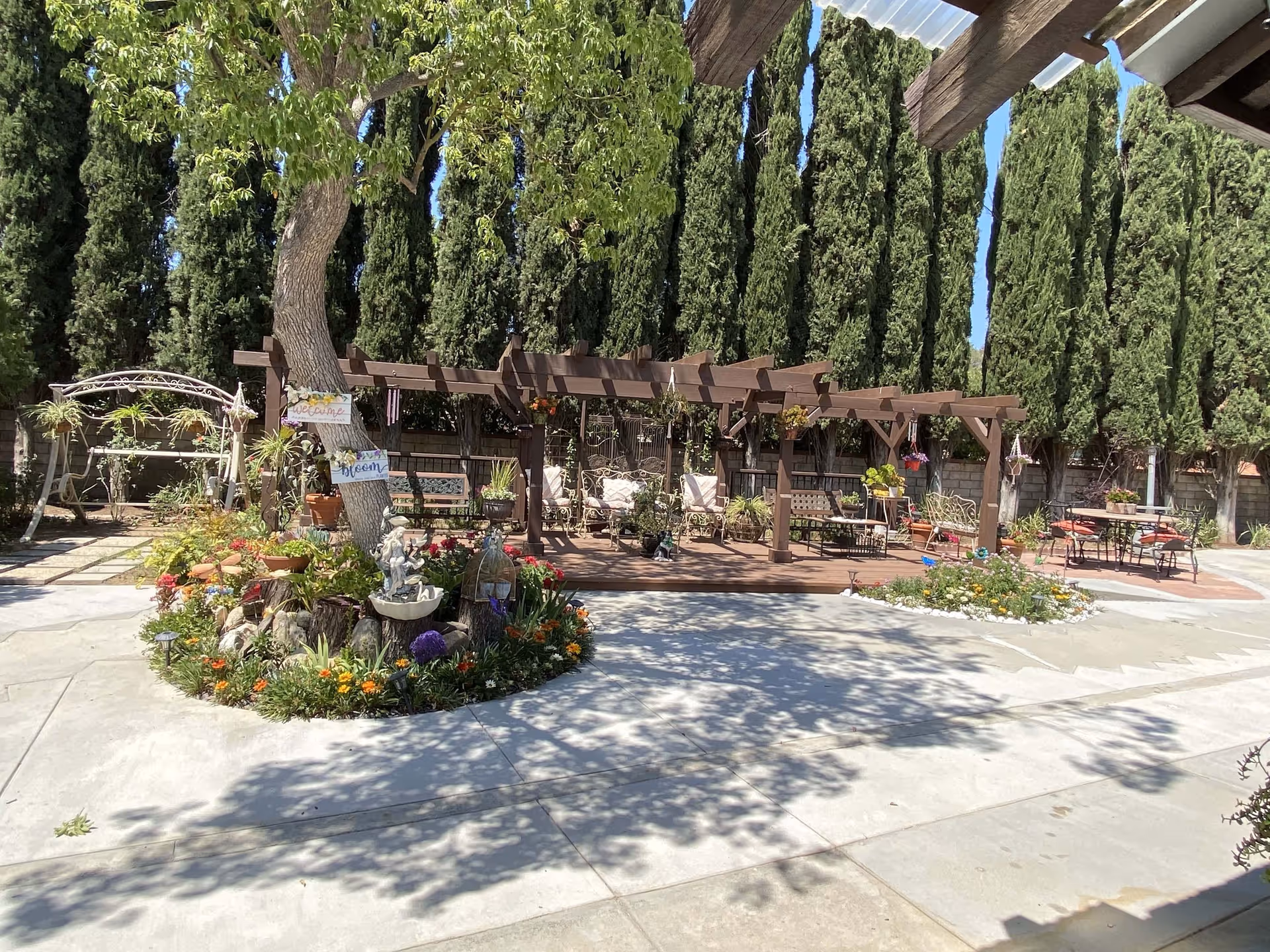 Outdoor patio area at Cypress Residence featuring a wooden pergola with hanging plants and cushioned chairs. There is a circular flower bed with various colorful flowers and garden decorations surrounding a tree with welcome signs. Tall evergreen trees line the background, and there are additional tables and chairs on the paved patio.