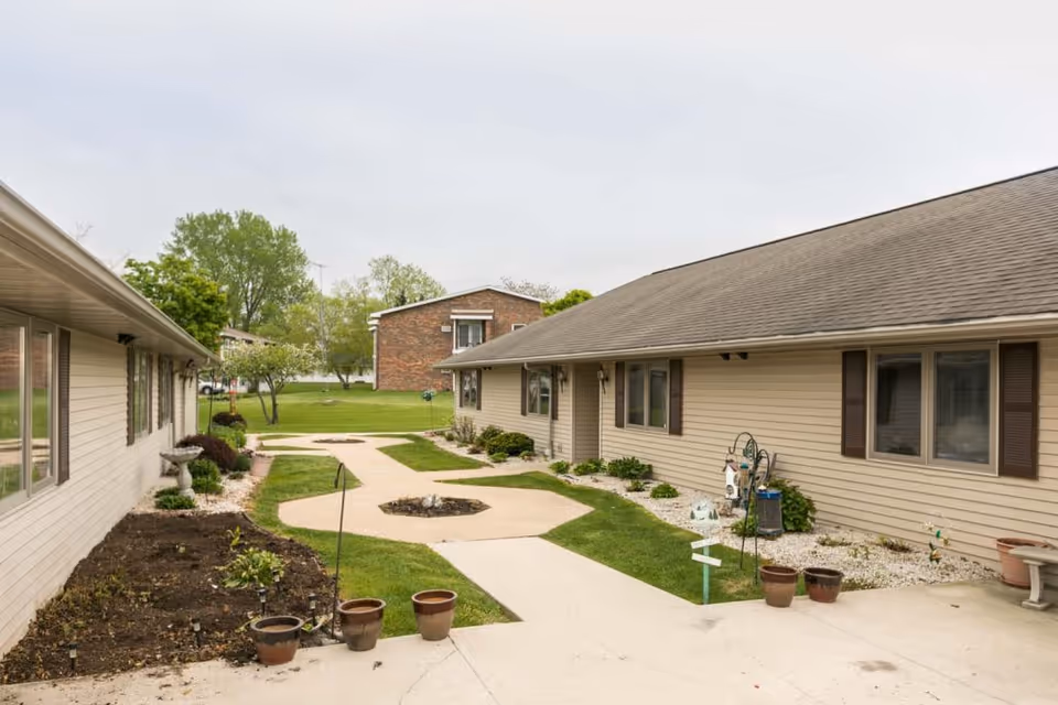 Outdoor courtyard area between two single-story beige buildings with brown shutters, featuring a concrete walkway, small garden beds, potted plants, and green grass under an overcast sky.