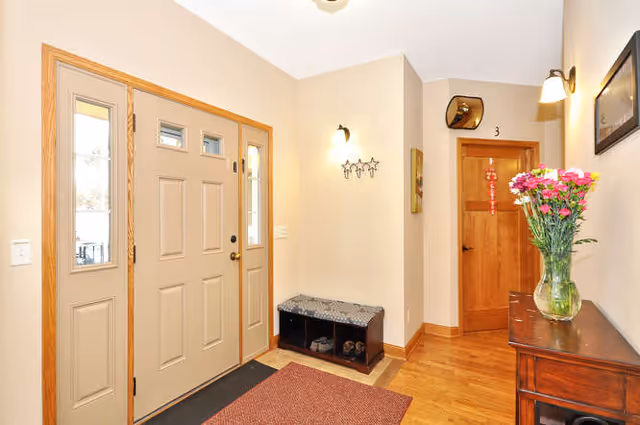 Entryway of a home with a beige front door featuring glass panels on each side. There is a red rug on the wooden floor, a small bench with storage underneath, and a wooden cabinet with a vase of pink and white flowers on top. The walls are light-colored with wall-mounted lights and some decorative items, including a star-shaped wall hook and a framed picture. A wooden door marked with the number 3 is visible in the background.