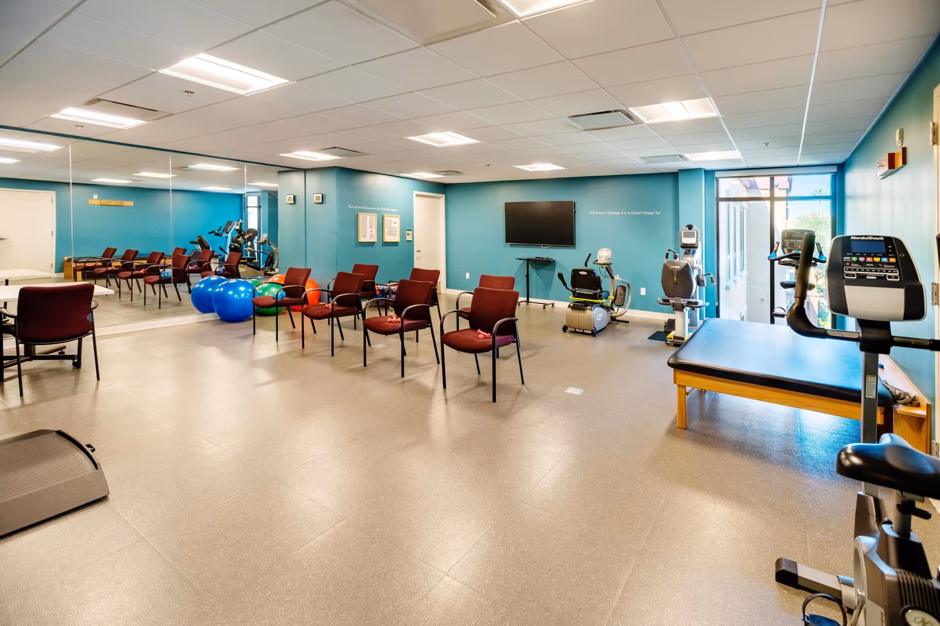 A spacious fitness and rehabilitation room with exercise equipment including stationary bikes, a treadmill, and therapy balls. Several chairs are arranged in rows facing a wall-mounted TV. The walls are painted blue, and one side of the room has large mirrors.