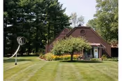 Brown triangular-roofed building with a small tree, modern lawn sculpture, and neatly mowed grass surrounded by tall trees.