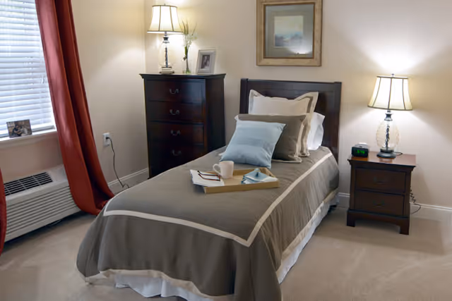 Neatly made single bed in a cozy bedroom with a nightstand, dresser, lamps, and a window with red curtains.