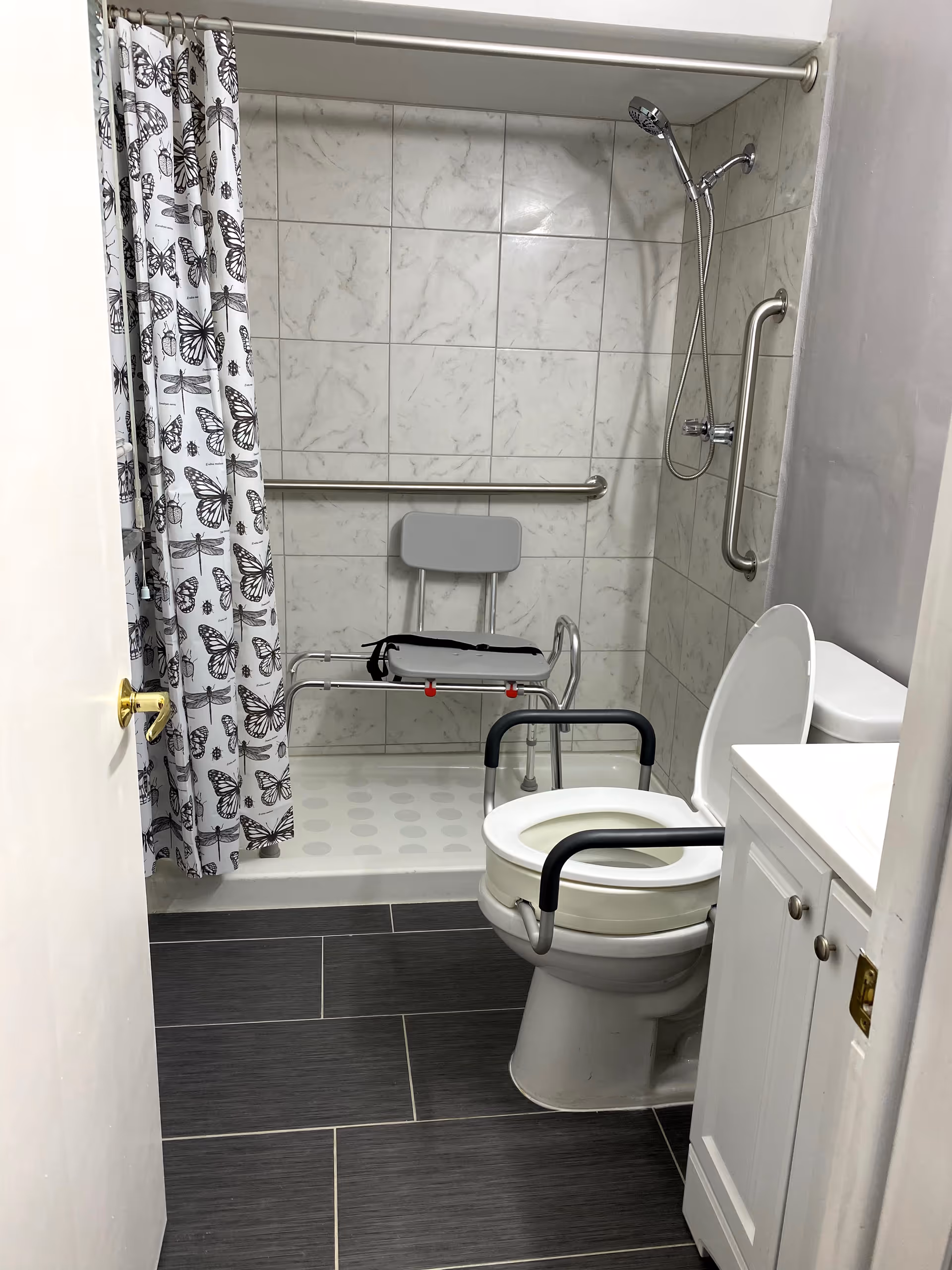 A bathroom with a walk-in shower featuring a butterfly-patterned shower curtain, a shower chair, and grab bars on the wall. There is a toilet with arm supports and a raised seat, and a white vanity with a sink. The floor is covered with dark gray tiles.