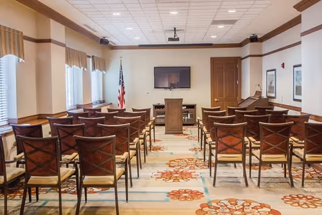 A meeting or presentation room with rows of wooden chairs facing a podium and a wall-mounted flat screen TV. The room has beige walls, a patterned carpet, several windows with blinds and valances, an American flag in the corner, and framed artwork on the walls.