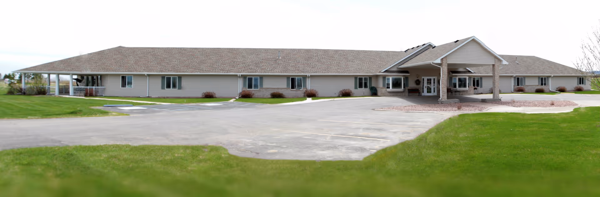 Single-story building with a covered entrance and multiple windows, surrounded by a green lawn and a paved parking area, under a cloudy sky.