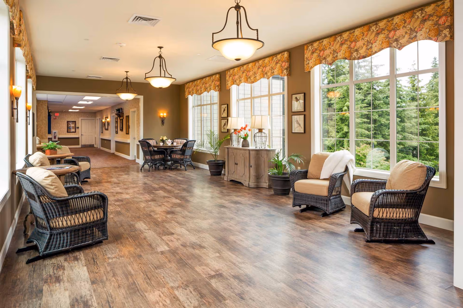 A spacious, well-lit common area in a senior living facility with large windows showing green trees outside. The room features wicker chairs with cushions, small tables, a wooden sideboard with lamps and plants, and a dining table with chairs. The floor is wood-style, and the walls are painted in warm tones with floral valances on the windows and ceiling light fixtures.