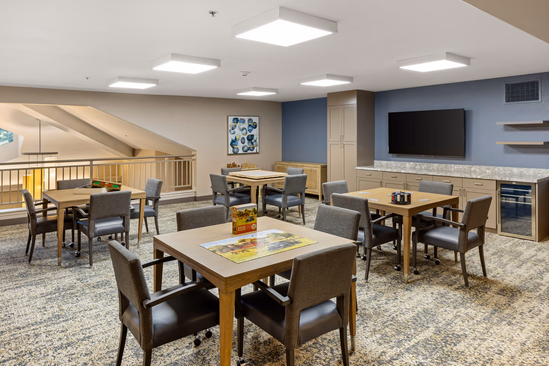 A well-lit common room with several wooden tables and cushioned chairs arranged for group activities or dining. The room features a carpeted floor, a large flat-screen TV mounted on a blue accent wall, and cabinets with a countertop below the TV. There are puzzles and games on the tables, and a railing overlooks a lower level.