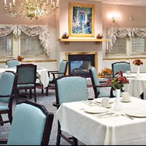 A dining room with tables covered in white tablecloths, each set with plates, cups, and silverware. The chairs have light blue upholstery with dark wooden frames. There are floral centerpieces on the tables. The room features a fireplace with a painting above it, two windows with floral curtains, and a chandelier hanging from the ceiling.