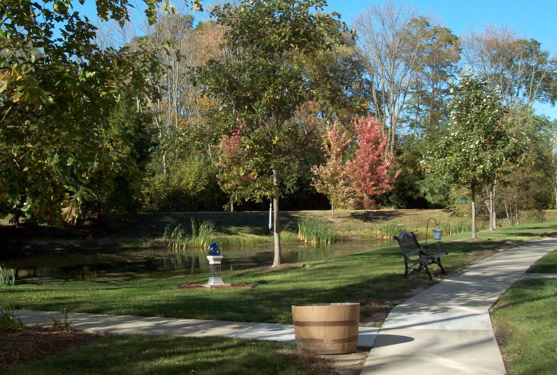A peaceful outdoor scene at Lakewood Tower Apartments featuring a curved concrete walkway, a wooden barrel planter, a black metal bench, and a small pond surrounded by green grass and trees with some autumn-colored leaves under a clear blue sky.