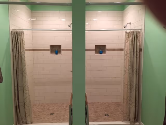 Two adjacent shower stalls with white tiled walls and beige tiled floors, each with a shower curtain and a small recessed shelf holding a blue bottle. The walls around the showers are painted light green.