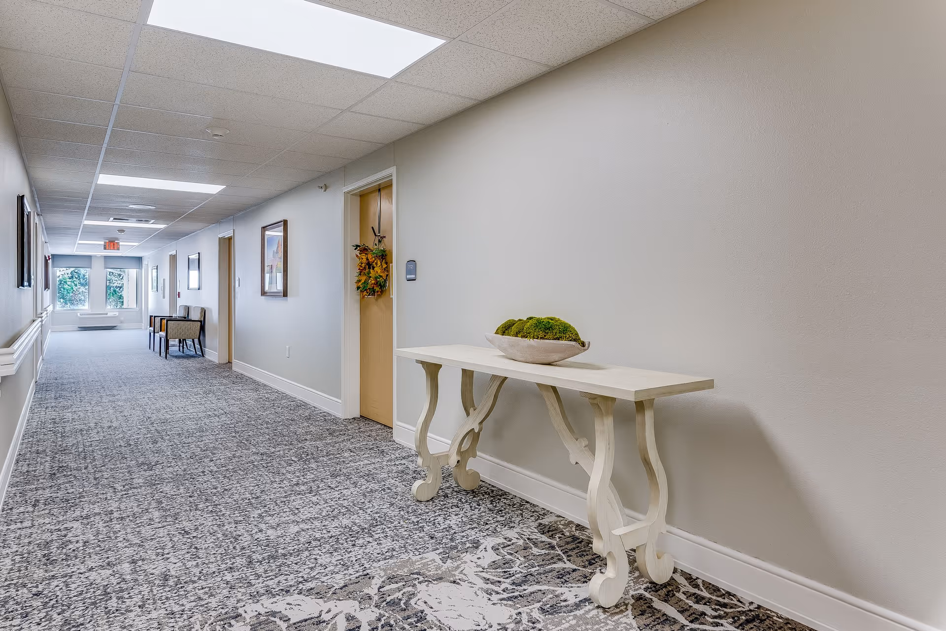 A long, well-lit hallway in a senior living facility with light-colored walls and patterned carpet. There is a white decorative table with a bowl of green moss on the right side, and several doors and framed pictures along the left wall. At the end of the hallway, there are windows letting in natural light and a couple of chairs placed against the wall.