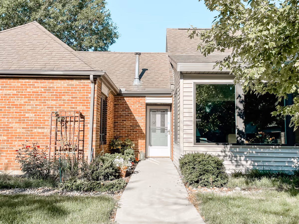 Front entrance of a single-story brick and siding building with a concrete walkway, shrubs, and a large window.