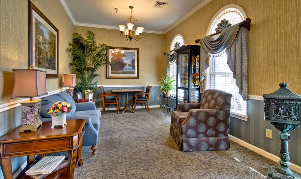 Bright, traditionally furnished sitting room with upholstered armchairs and a sofa, side tables and lamps, a small table with chairs, and decorative arched windows.