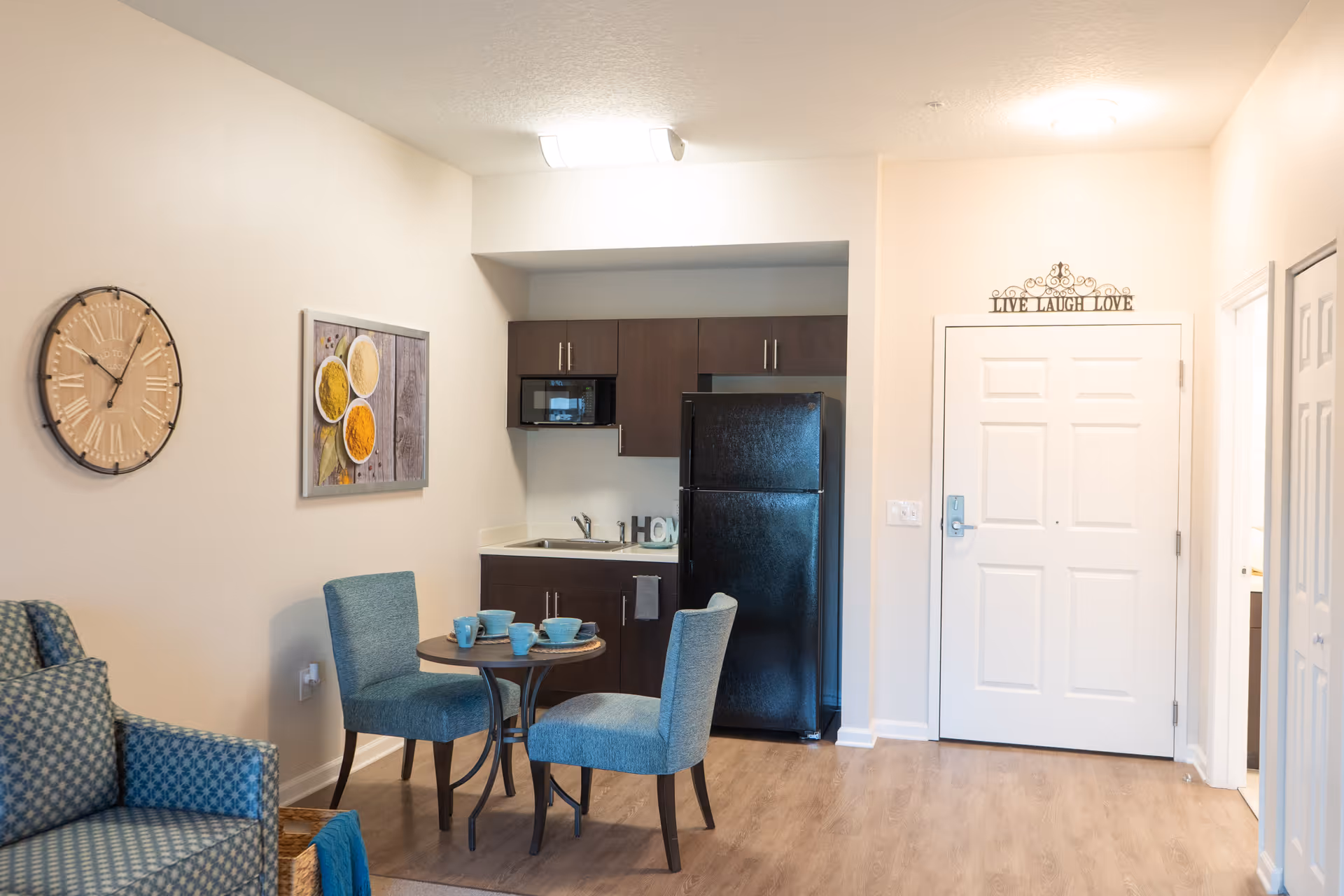 A small kitchen and dining area in a senior living facility apartment. The kitchen has dark brown cabinets, a black refrigerator, a microwave, and a sink. In front of the kitchen is a round table with two blue upholstered chairs and blue dishware on the table. To the left is a blue patterned armchair. On the wall above the armchair is a large round clock and a framed picture of spices. The entrance door is white with a decorative sign above it that reads 'LIVE LAUGH LOVE'. The floor is light wood, and the walls are painted white.