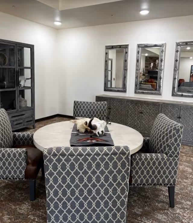 A dining area with a round table surrounded by four patterned upholstered chairs. The table has a decorative runner and a centerpiece featuring animal skulls. Behind the table is a sideboard with intricate designs and three large decorative mirrors hanging above it. To the left, there is a dark wooden cabinet with glass doors displaying decorative items. The room has a neutral color palette with white walls and a patterned carpet.