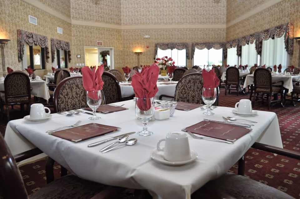 A formal dining room with multiple tables set for a meal. Each table is covered with a white tablecloth and has maroon cloth napkins folded in glasses, white cups and saucers, silverware, and menus. The room has patterned wallpaper, large windows with valances, and carpeted floors with a red and gold pattern.