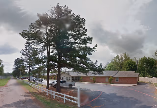 Exterior view of a single-story brick building with a parking lot in front, surrounded by trees and a white fence along the road. Several vehicles are parked near the building under a cloudy sky.