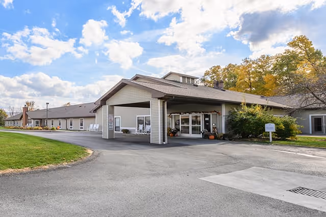 Exterior view of a single-story senior living facility building with a covered entrance driveway, surrounded by green grass and trees under a partly cloudy sky.