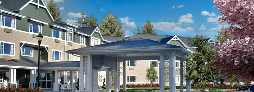Exterior view of a multi-story assisted living facility building with beige siding and white trim, featuring a covered entrance with white pillars, surrounded by landscaped greenery and flowering trees under a blue sky with some clouds.
