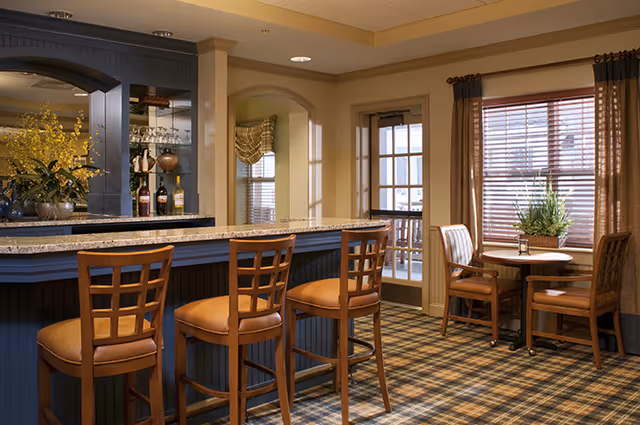 Interior view of a senior living facility lounge area featuring a bar with four wooden bar stools with cushioned seats, a granite countertop, and shelves with bottles and glassware behind the bar. To the right, there is a small round table with two wooden chairs near a window with brown curtains and blinds. The room has warm lighting and a patterned carpet.