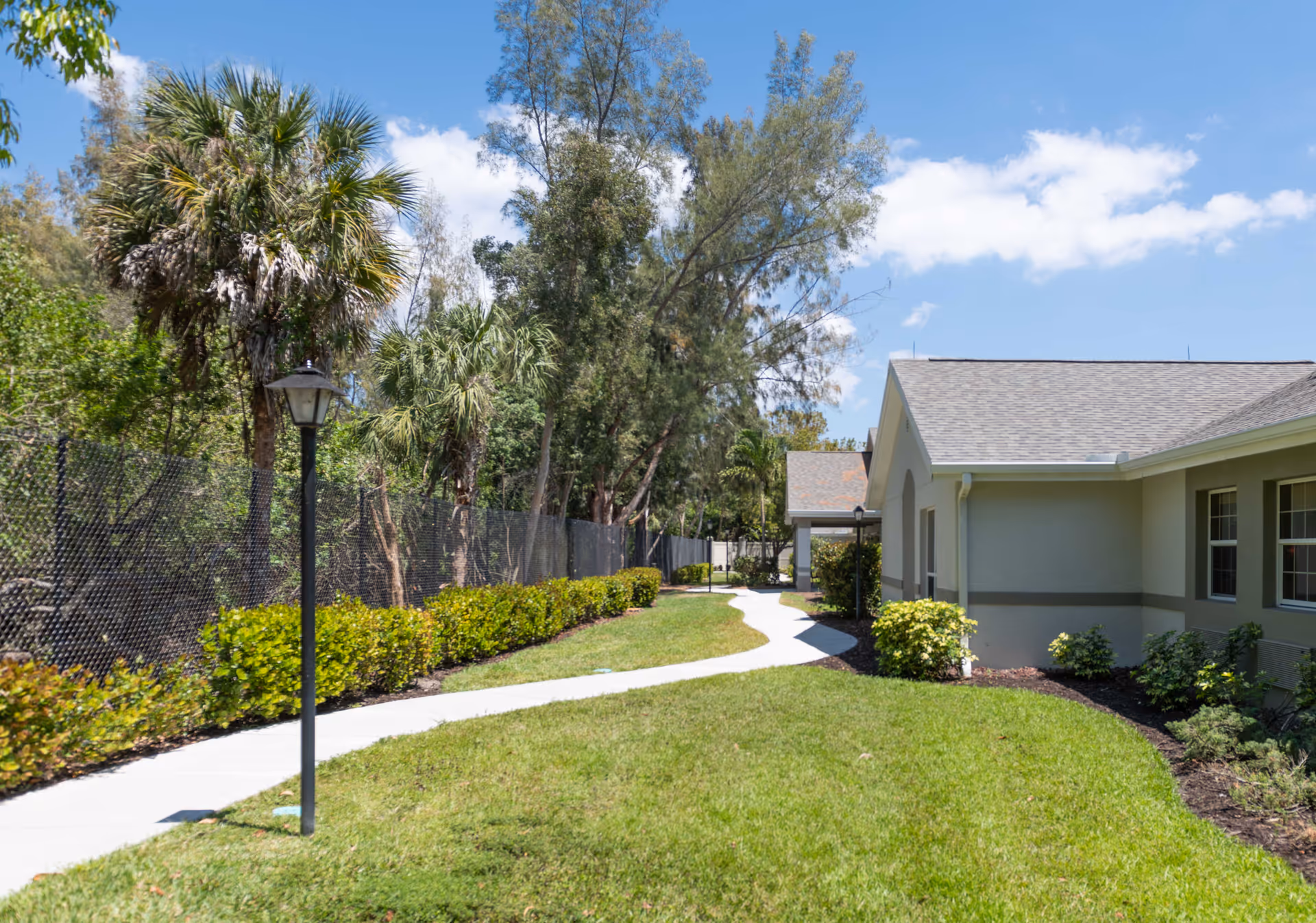 A sunny outdoor pathway winding through a garden area with green grass, bushes, palm trees, and other trees. A beige building with multiple windows is visible on the right side, and a black chain-link fence runs along the left side of the path. A black lamp post stands near the path.