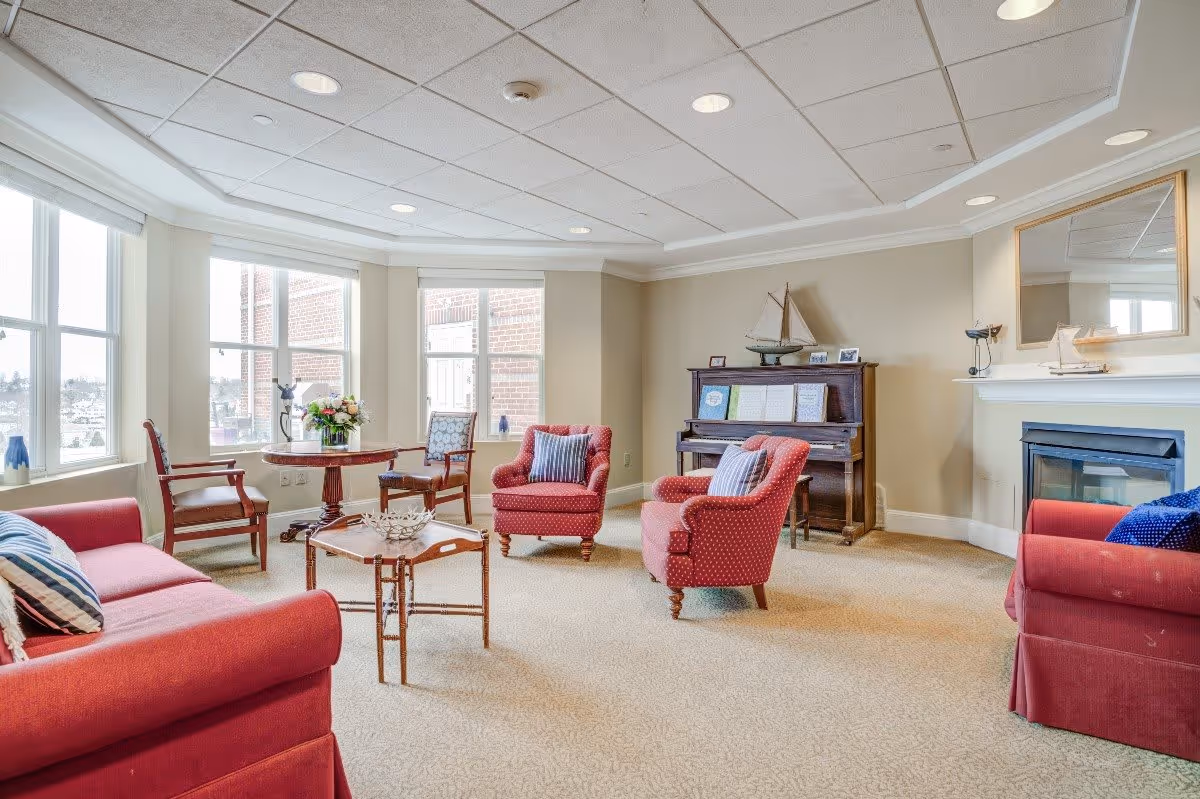 A bright and cozy living room with large windows letting in natural light. The room features red upholstered armchairs and sofas with striped pillows, a wooden coffee table, a round side table with a flower arrangement, a piano with decorative items on top, and a fireplace with a mirror above it.