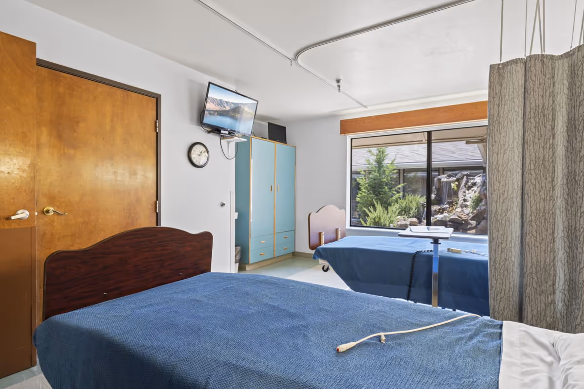A nursing facility room with two beds covered in blue blankets, a wooden door, a wall-mounted TV, a clock, a blue wardrobe, and a large window showing greenery outside. A privacy curtain is partially visible on the right side.