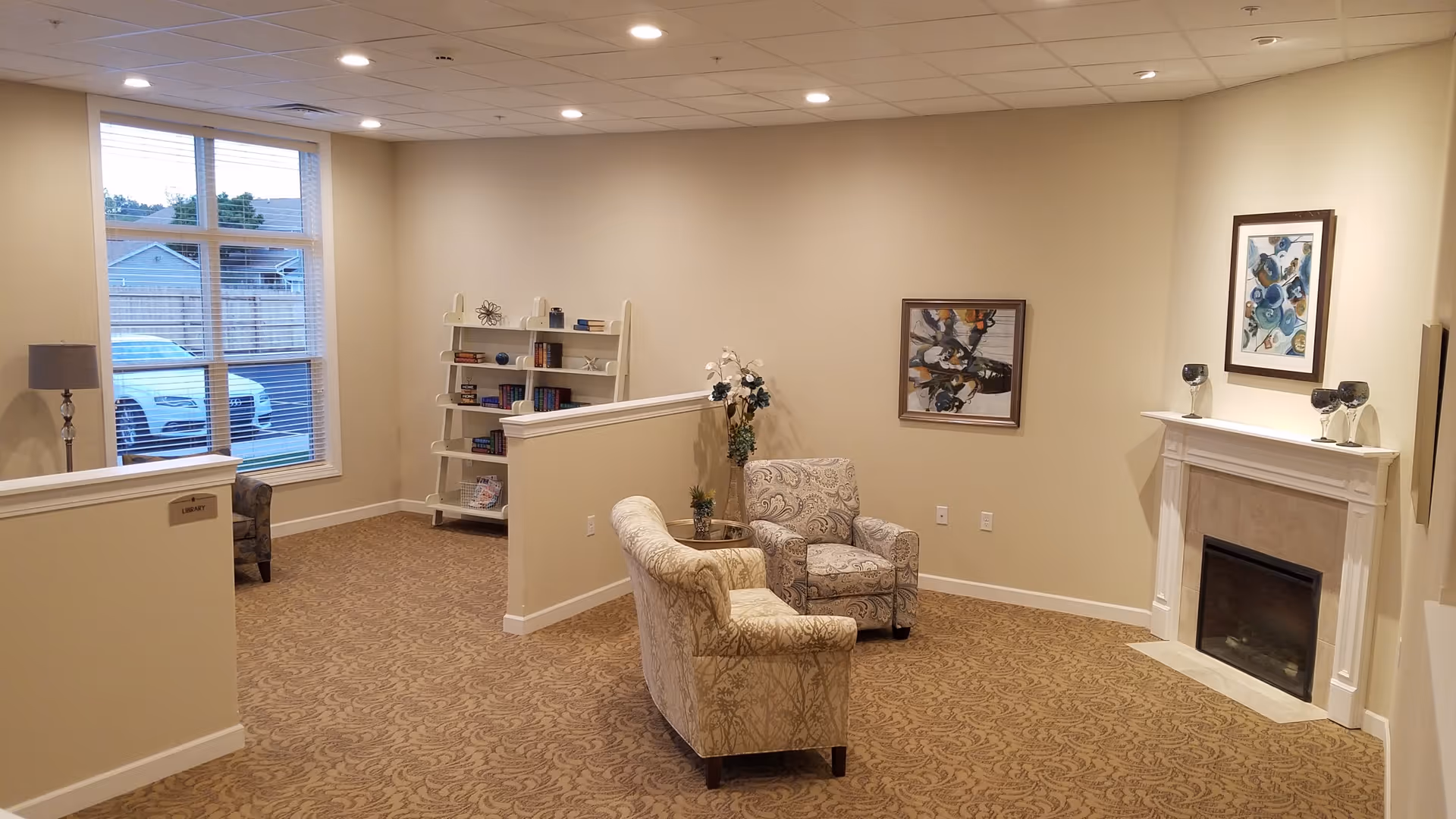 A cozy living room area with two patterned armchairs facing each other near a white fireplace. The room has beige walls and carpet with a subtle pattern. There are two framed abstract paintings on the walls, a white bookshelf with books and decorative items, a floor lamp near a large window showing a parked car outside, and a small table with a flower arrangement between the chairs.