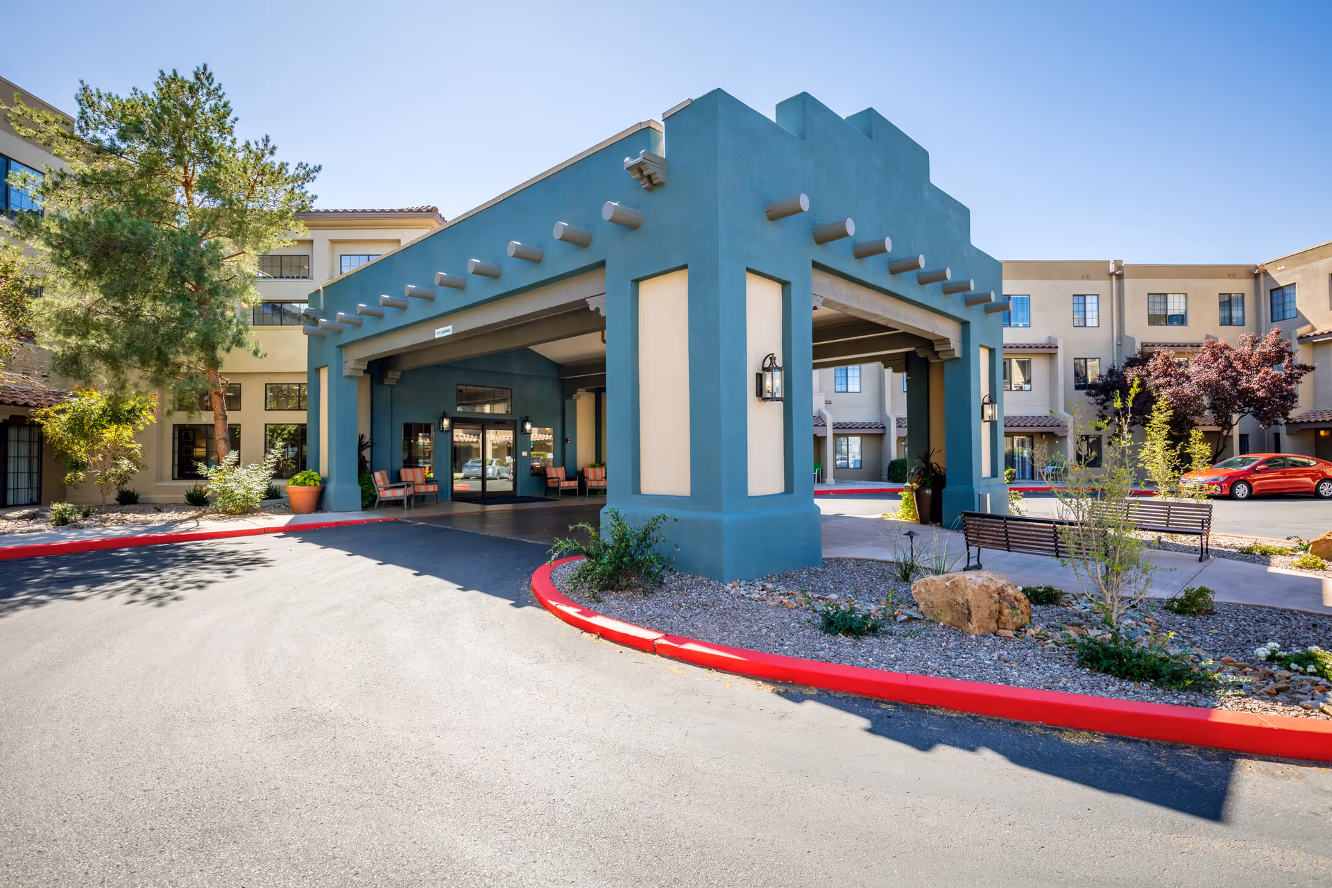 Entrance area of Fairwinds - Rio Rancho senior living facility featuring a covered drop-off zone with teal and beige pillars, outdoor benches, landscaped plants, and a multi-story building in the background under a clear blue sky.