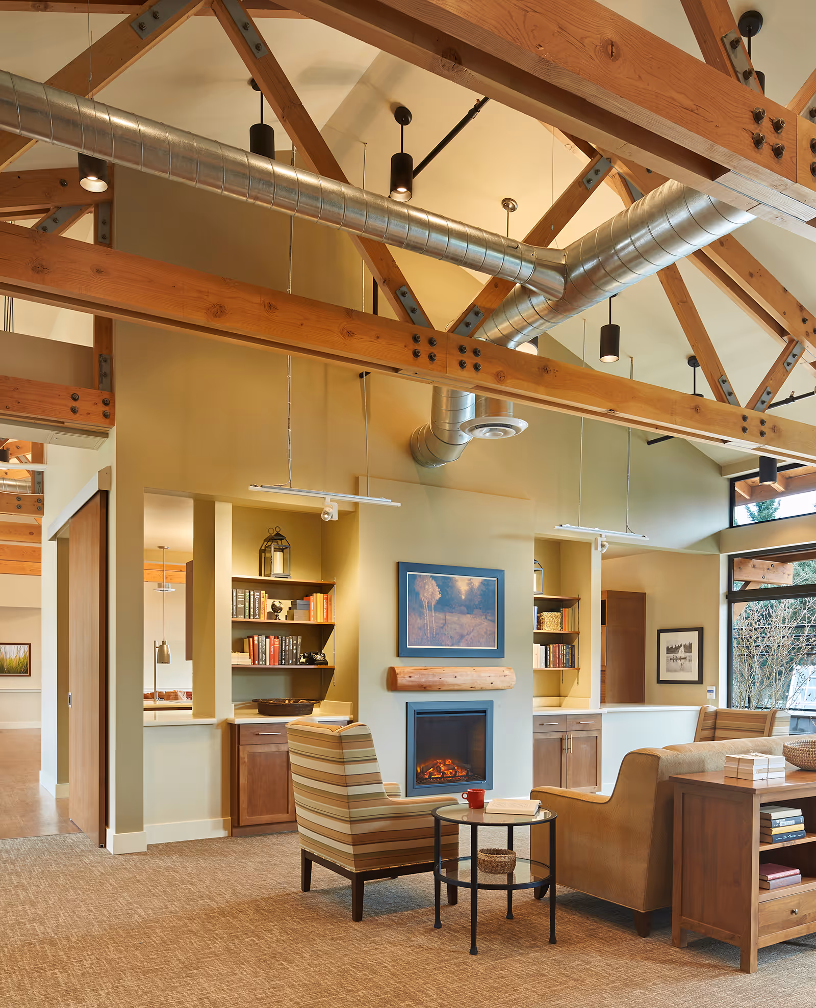 A cozy living room area with exposed wooden beams and metal ductwork on the ceiling. The room features a fireplace with a wooden mantel, a framed landscape painting above it, built-in shelves with books and decorative items, a striped armchair, a beige sofa, and a small round table with a red mug and an open book. Large windows allow natural light to fill the space.