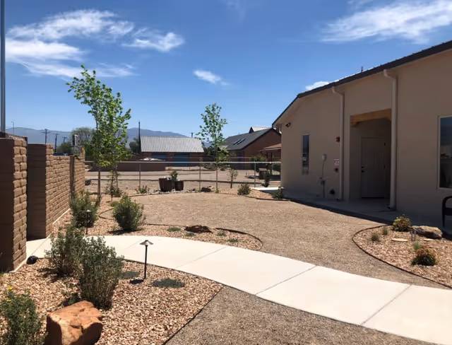 Sunny landscaped courtyard with a curved concrete walkway, gravel beds, small shrubs and a single-story building under a blue sky.