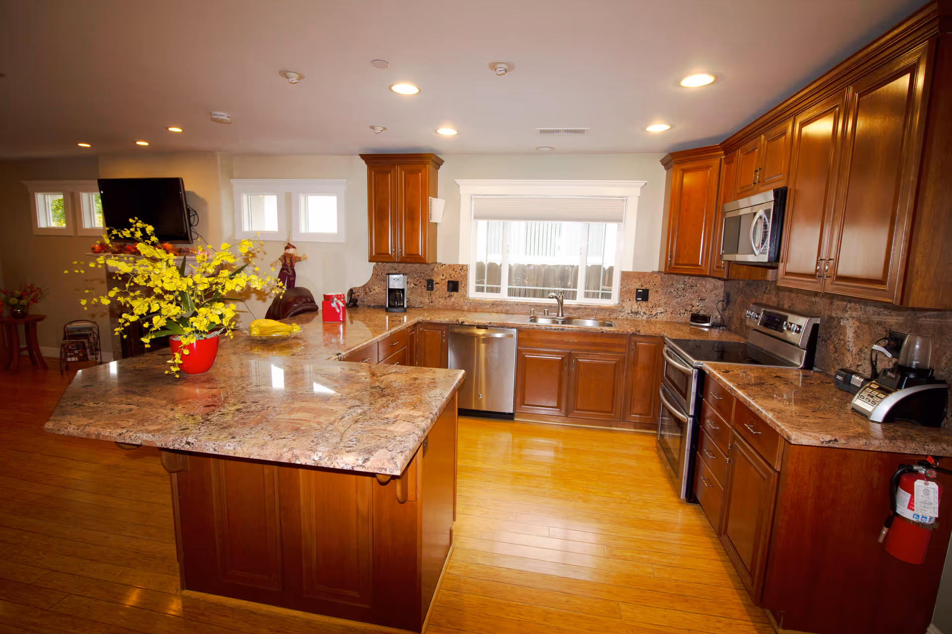 A spacious kitchen with wooden cabinets and granite countertops. The kitchen features a large island with a red vase holding yellow flowers, stainless steel appliances including a dishwasher, oven, and microwave, and a window above the sink letting in natural light. The floor is made of light-colored wood, and there is a fire extinguisher mounted on the right side near the cabinets.