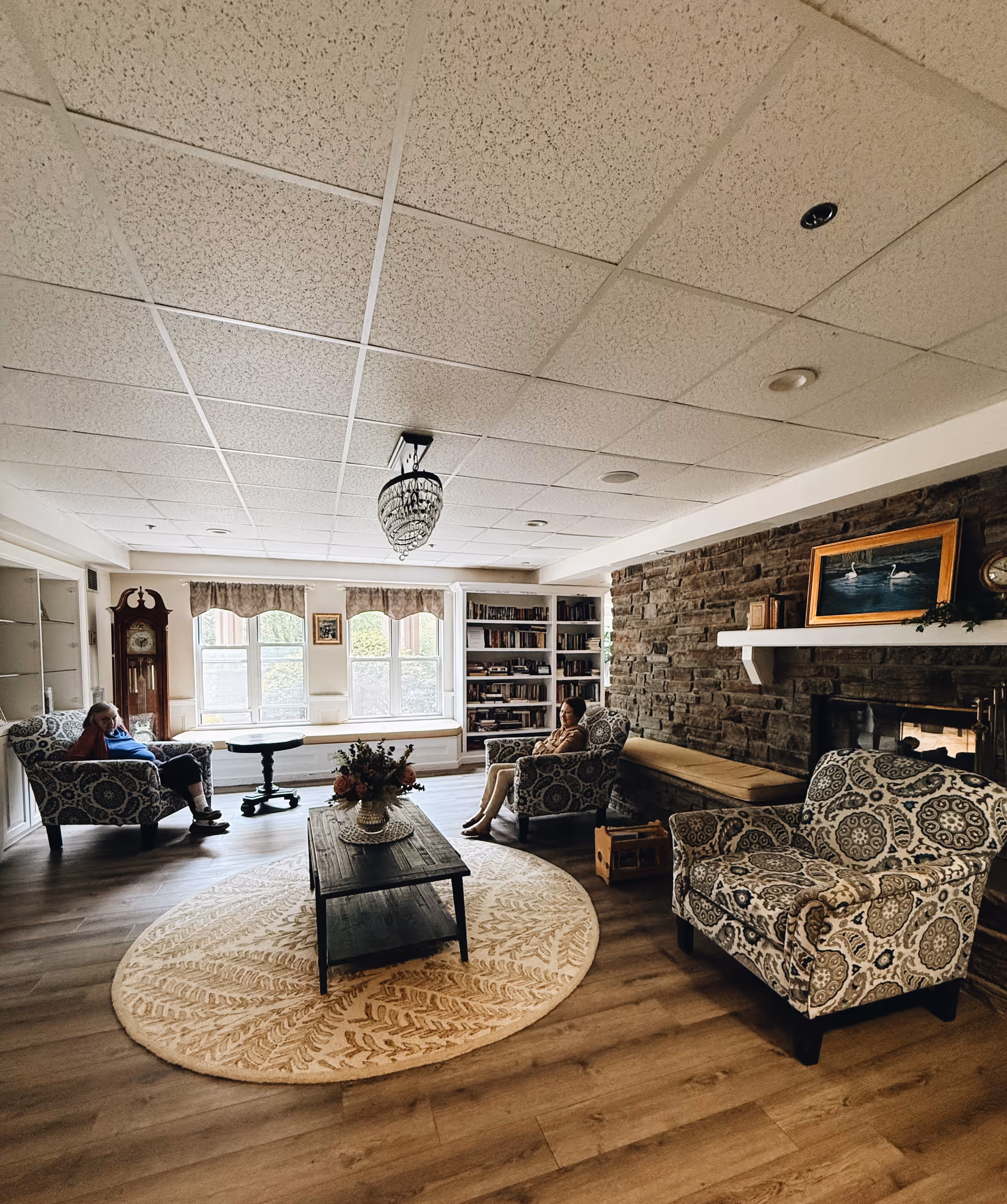 Cozy communal lounge with patterned armchairs, a round rug and coffee table, a stone fireplace, bookshelves and two people seated near windows.
