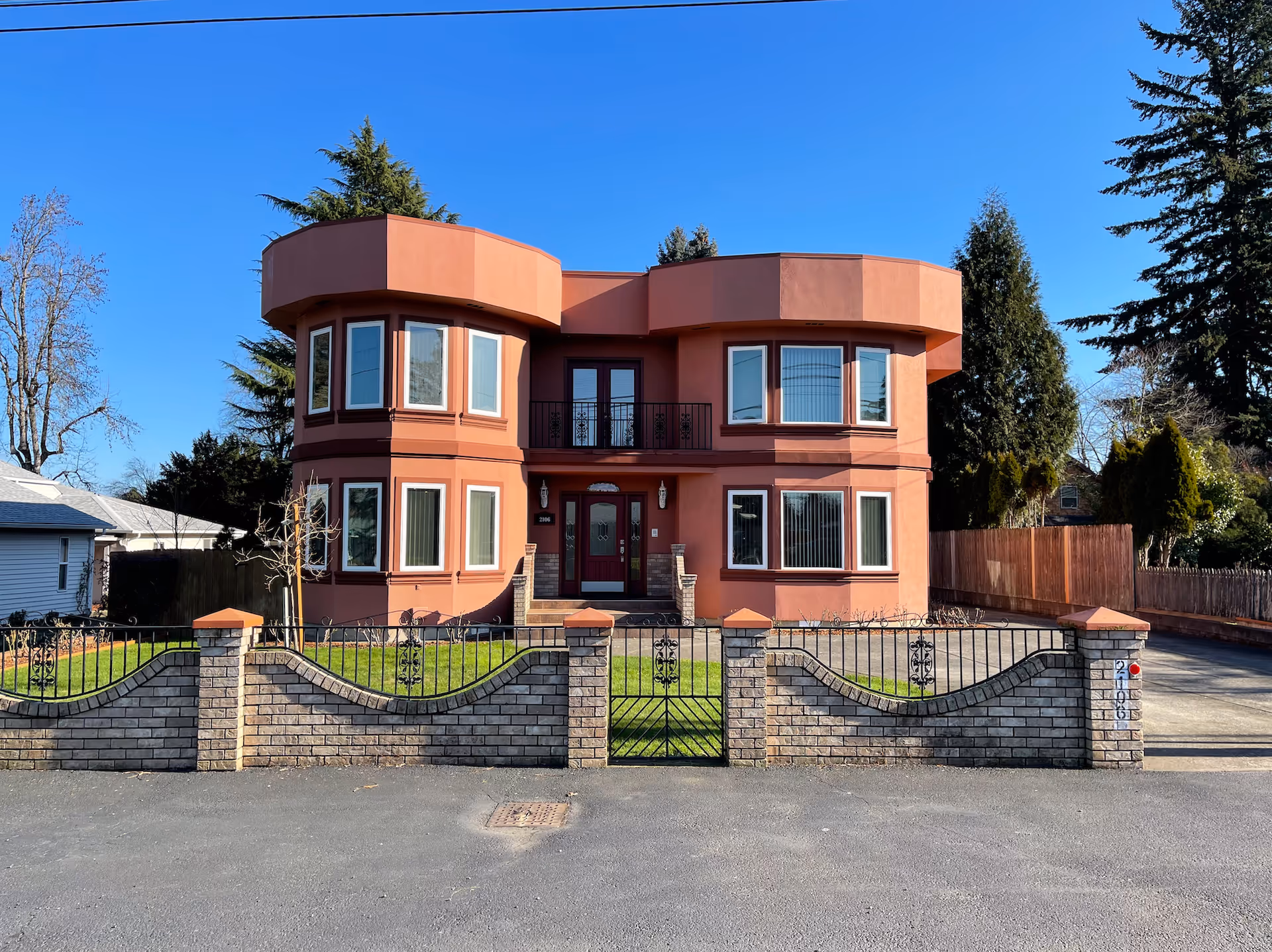 Front exterior view of a two-story building with a reddish-brown facade, multiple windows, a central entrance with double doors, and a decorative brick and wrought iron fence enclosing a small lawn. Tall trees and a clear blue sky are visible in the background.