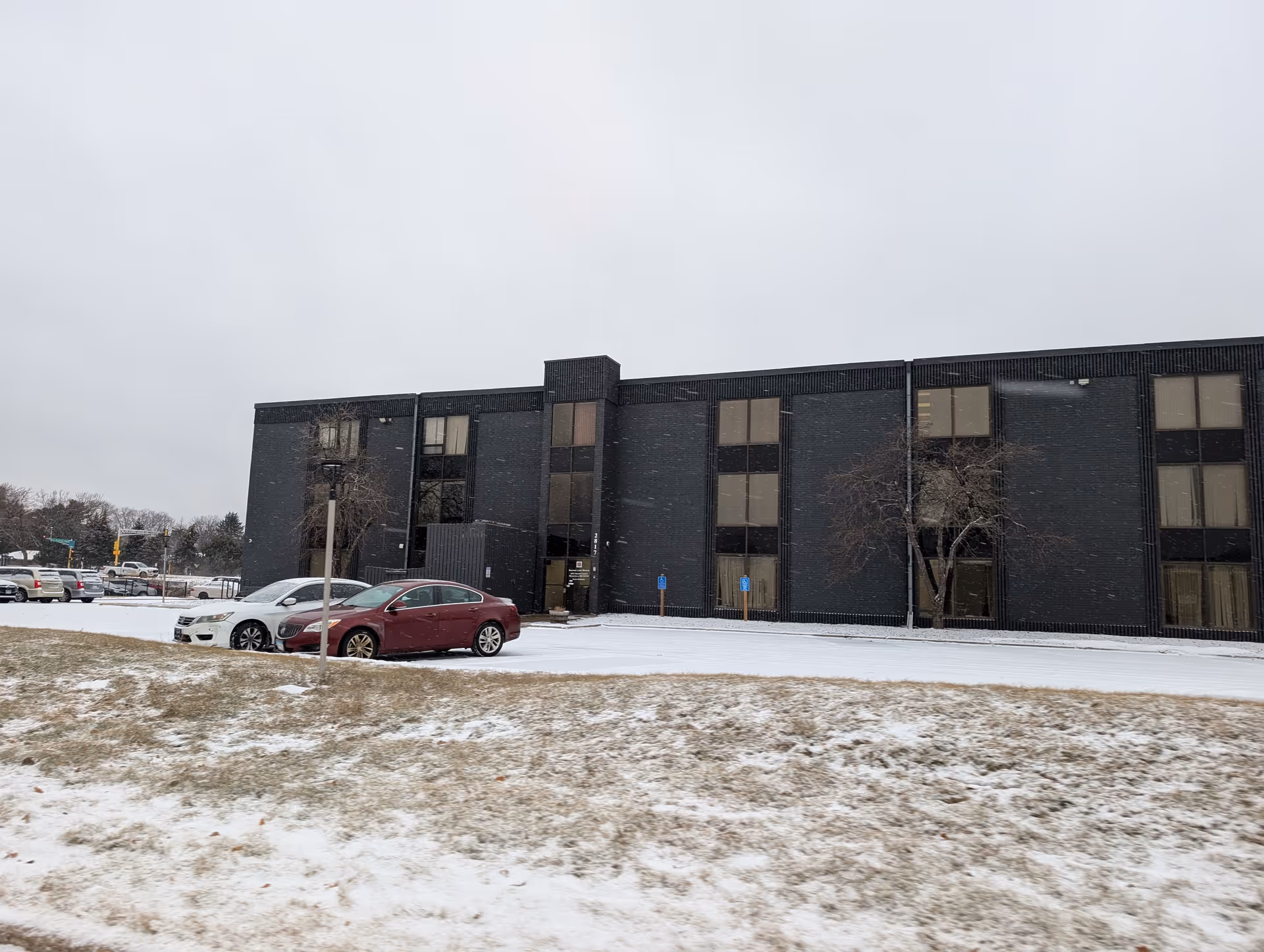 Snow-covered lawn and parking area with a few cars in front of a dark three-story commercial building.