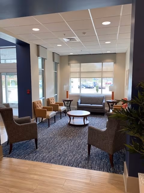 A cozy seating area in a senior living facility with a gray sofa, four armchairs in gray and tan colors, a round wooden coffee table in the center, two side tables with orange lamps, large windows with beige blinds, and a blue carpet on a wooden floor.