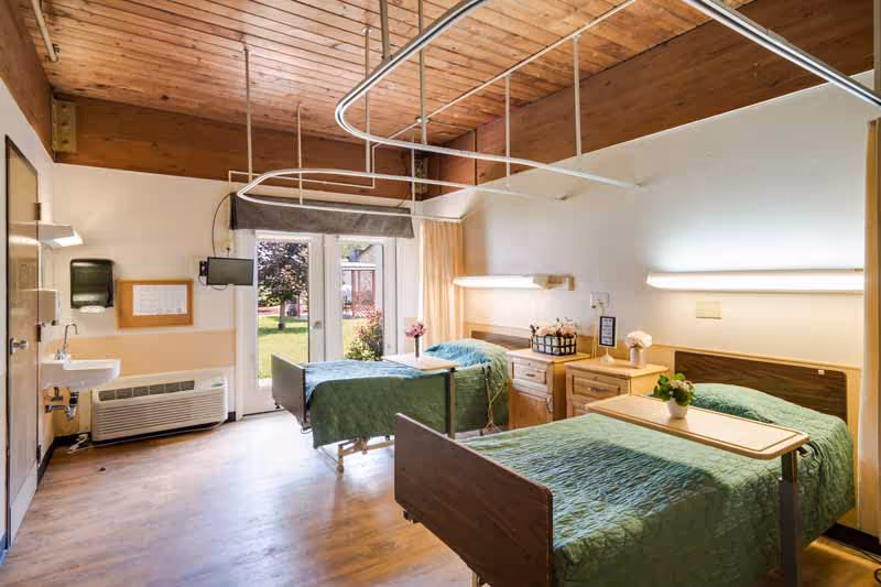 A bright and clean senior living bedroom with two hospital-style beds covered in green quilts. Each bed has a bedside table with flowers and a wall-mounted light above. There is a large window and glass door letting in natural light and showing a view of the outdoor garden. The room has wooden ceiling panels and light-colored walls and flooring.