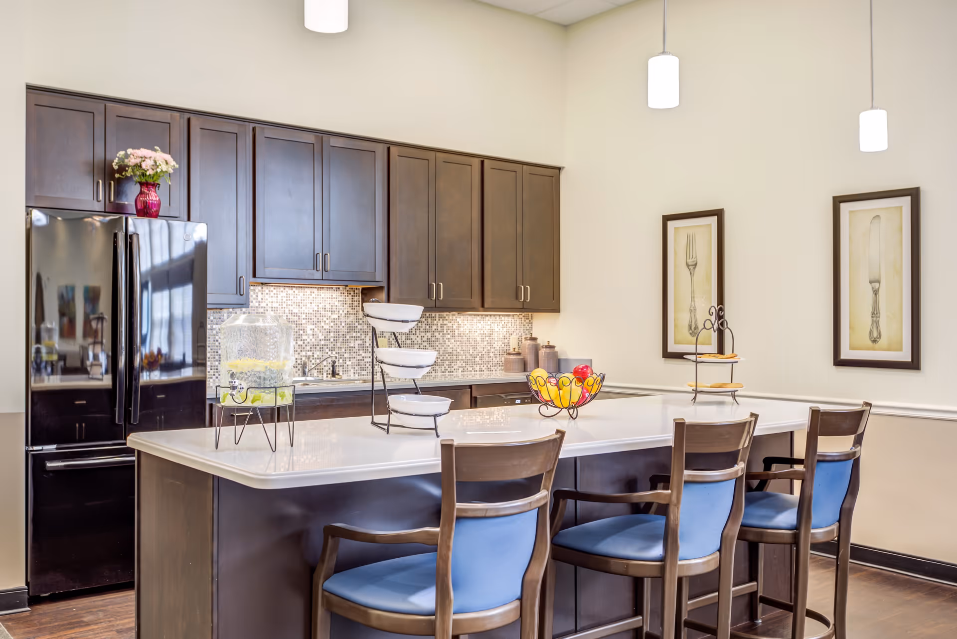 A modern kitchen area with dark wood cabinets, a black refrigerator, and a large white island countertop with four wooden chairs featuring blue cushions. The countertop has a glass beverage dispenser, a tiered serving tray, and a fruit bowl. Two framed pictures of a fork and knife hang on the wall, and three pendant lights hang from the ceiling.