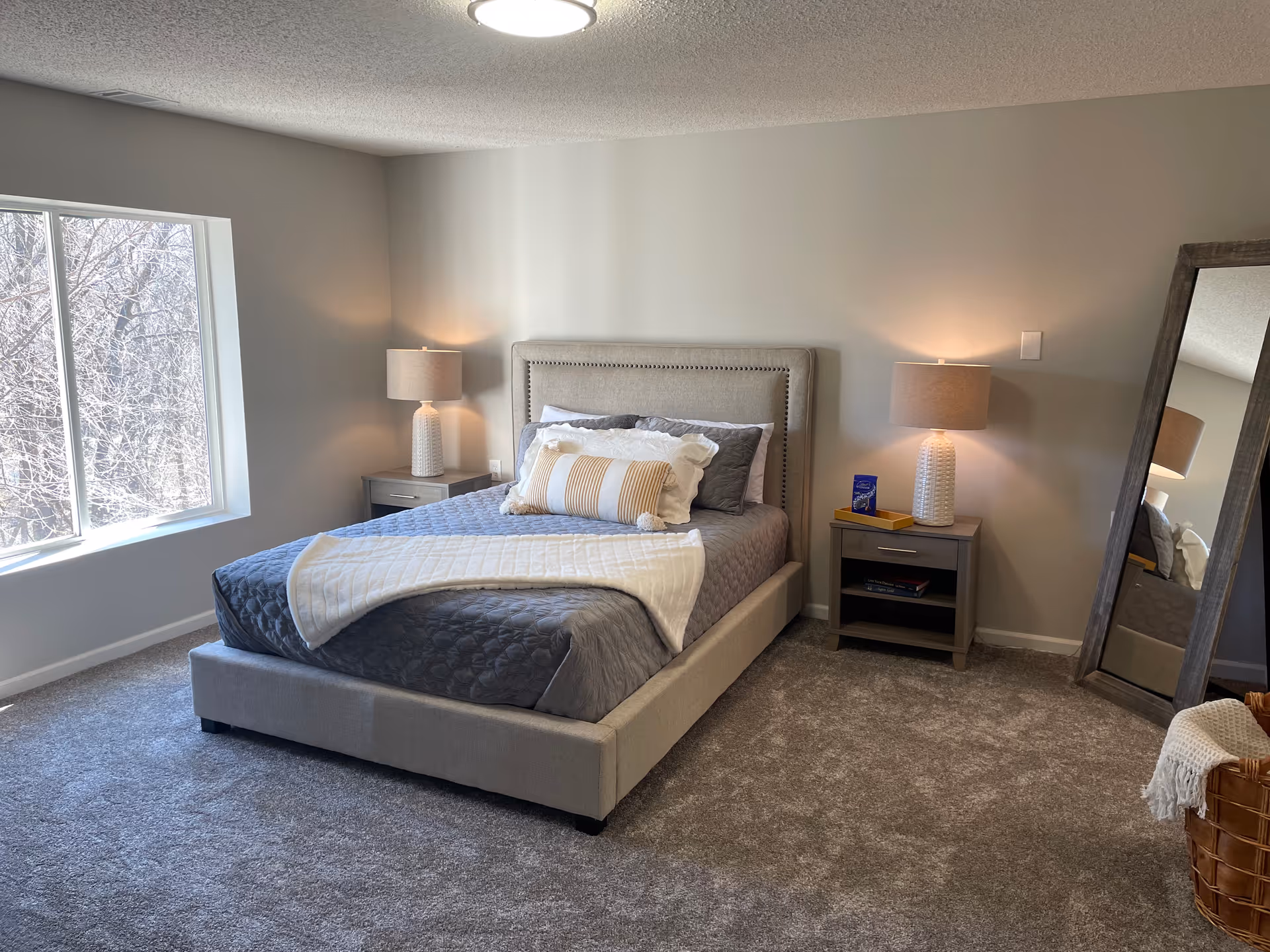 A neatly made bed with gray bedding and white pillows in a carpeted bedroom. Two matching bedside tables with lamps flank the bed. A large window on the left lets in natural light, and a tall standing mirror is positioned on the right side of the room next to a woven basket with a white throw.