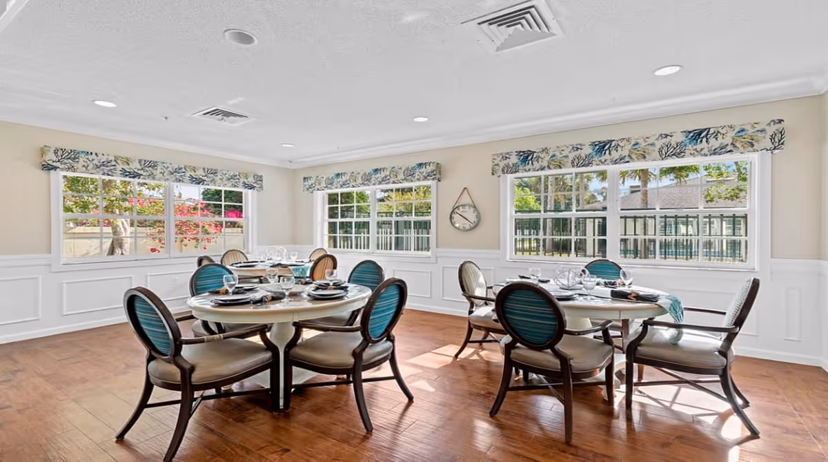Bright dining room with two round tables set for meals, each surrounded by six chairs with blue and beige upholstery. Large windows with floral valances let in natural light and offer views of greenery outside. The room has light-colored walls with white wainscoting and a wooden floor.