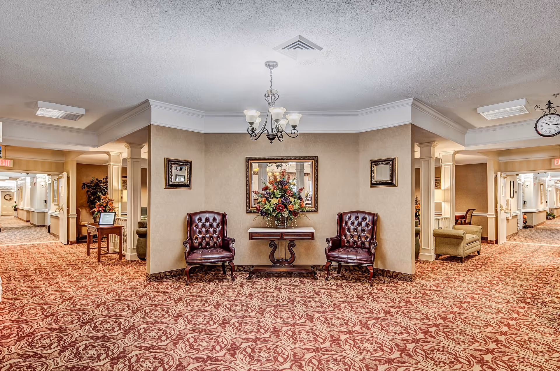 A spacious and well-lit interior hallway area with patterned carpet and beige walls. Two dark brown leather armchairs flank a small wooden table with a large floral arrangement. Above the table is a framed mirror. The hallway extends to the left and right with additional seating and decorative plants visible.