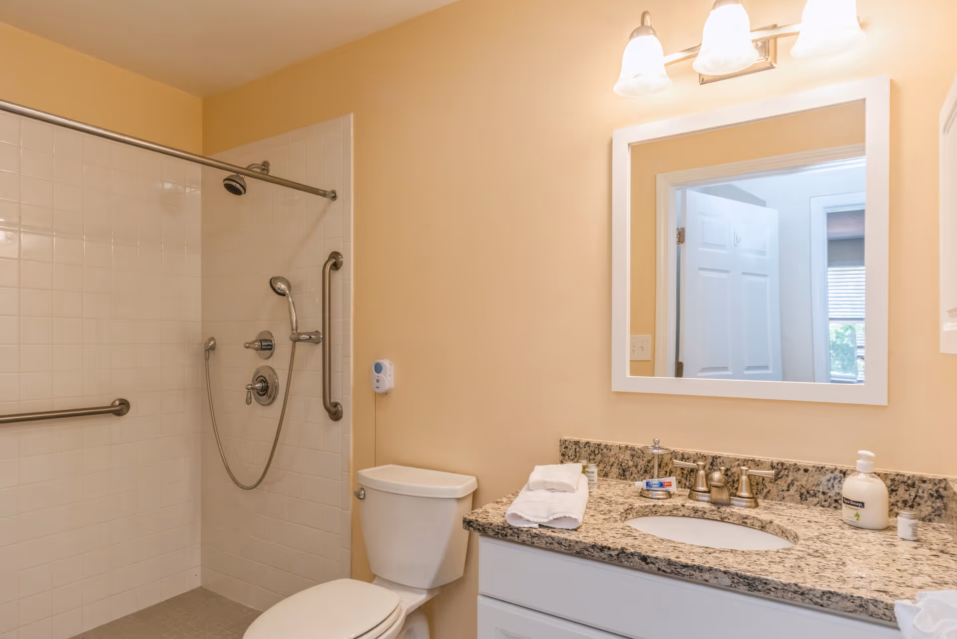 A bathroom with beige walls featuring a white toilet, a granite countertop with a sink, a large mirror above the sink, and a walk-in shower with white tiles and grab bars. The countertop has folded white towels, a soap dispenser, and some small toiletry items.