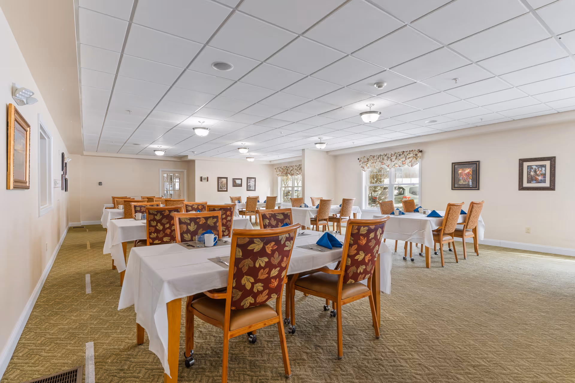 A dining room in a senior living facility with several tables covered in white tablecloths and set with blue napkins, cups, and placemats. The chairs have wooden frames with patterned upholstery featuring autumn leaves. The room has beige walls, carpeted floors, framed artwork on the walls, and windows with floral valances letting in natural light.