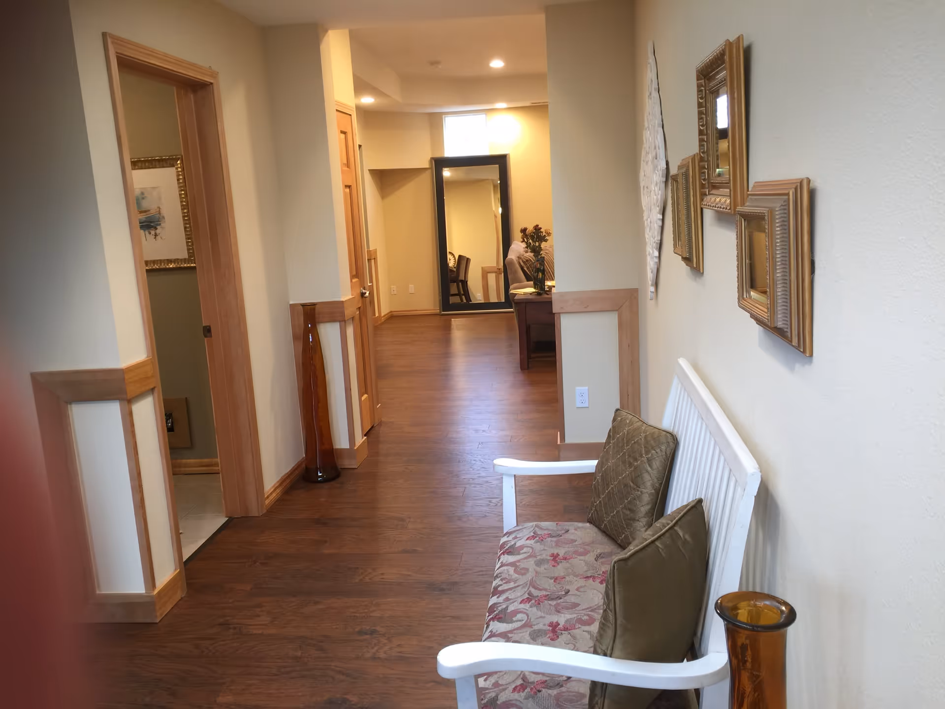 Interior hallway of a senior living facility with wooden flooring and beige walls. On the right side, there is a white bench with patterned cushions and decorative wall hangings above it. Further down the hallway, a large mirror reflects part of a sitting area with a table and chairs. There is a tall amber-colored vase on the floor near the bench and another similar vase near a doorway on the left.