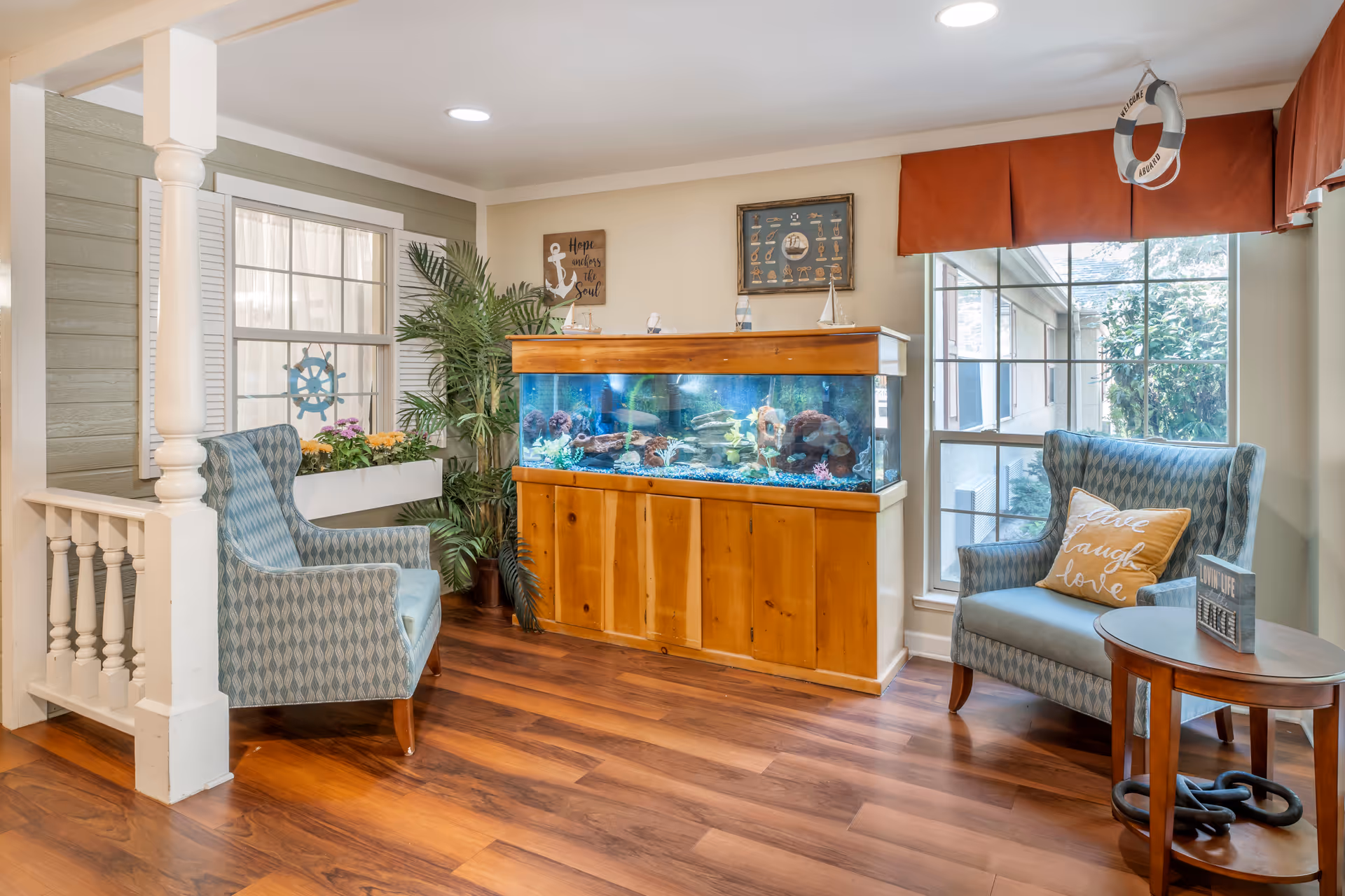 A cozy seating area in a senior living facility with two patterned armchairs, a wooden side table, and a large aquarium filled with colorful fish. The room features wooden flooring, a large window with red valance, decorative plants, and nautical-themed wall art including a life preserver and a sign that reads 'Hope anchors the soul.'