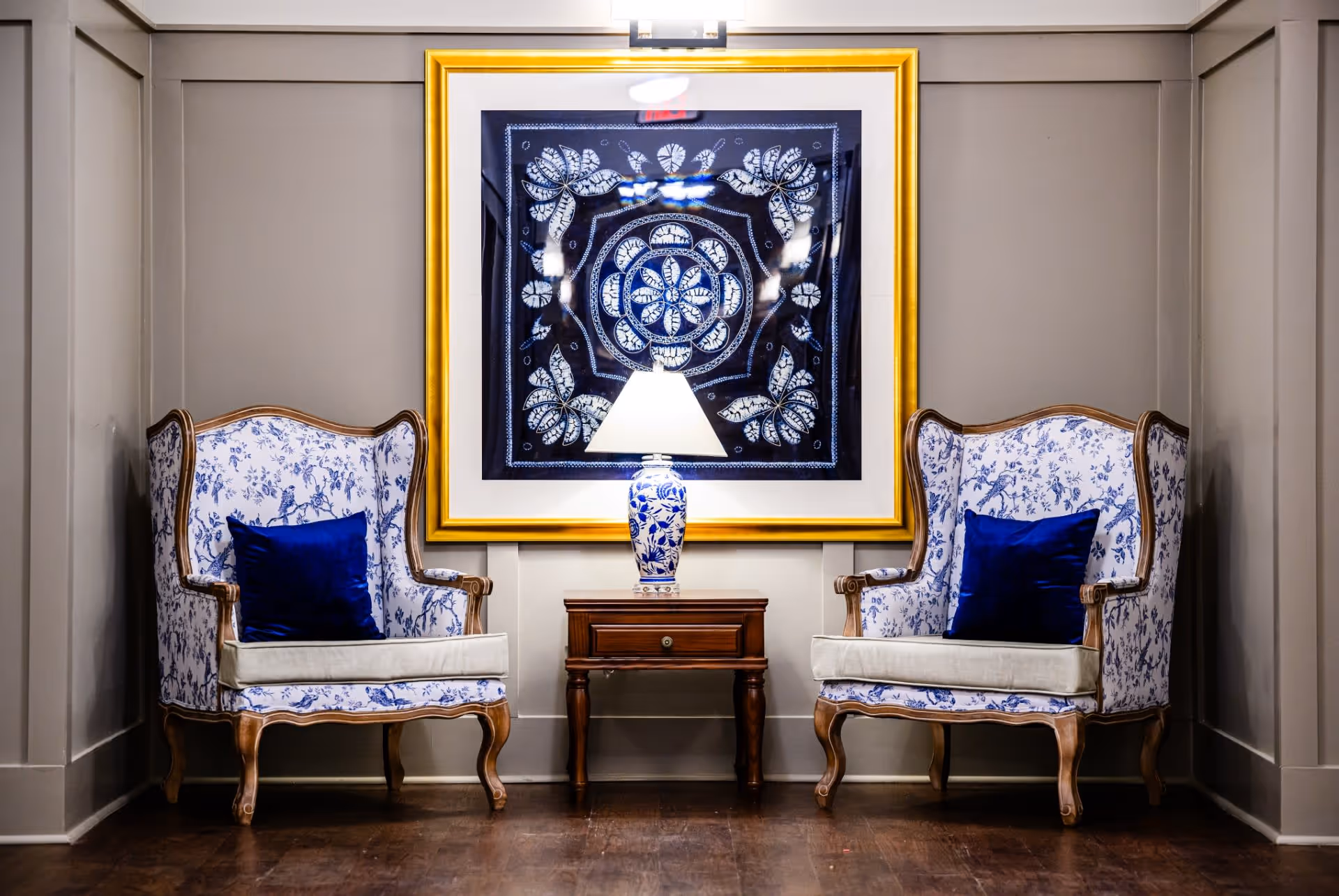Two blue-and-white upholstered armchairs flank a wooden side table with a blue-and-white lamp beneath a framed artwork on a paneled wall.