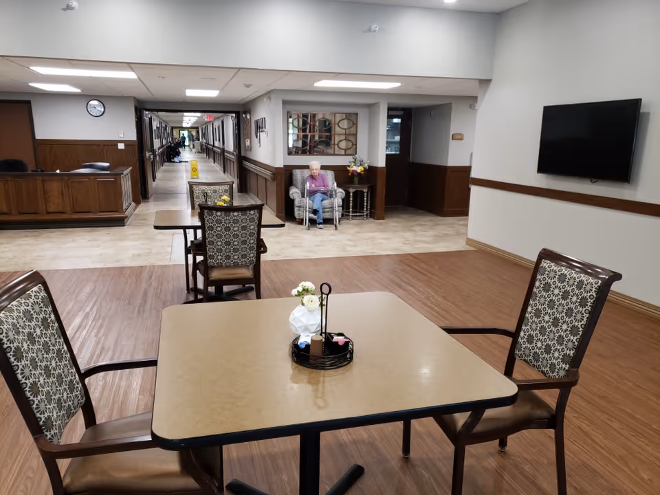 A seating area in a senior living facility with a table and four patterned chairs in the foreground. In the background, an elderly person is sitting in a cushioned chair near a small table with a flower arrangement. The area has wood and tile flooring, a wall-mounted TV, and a long hallway leading further into the facility.