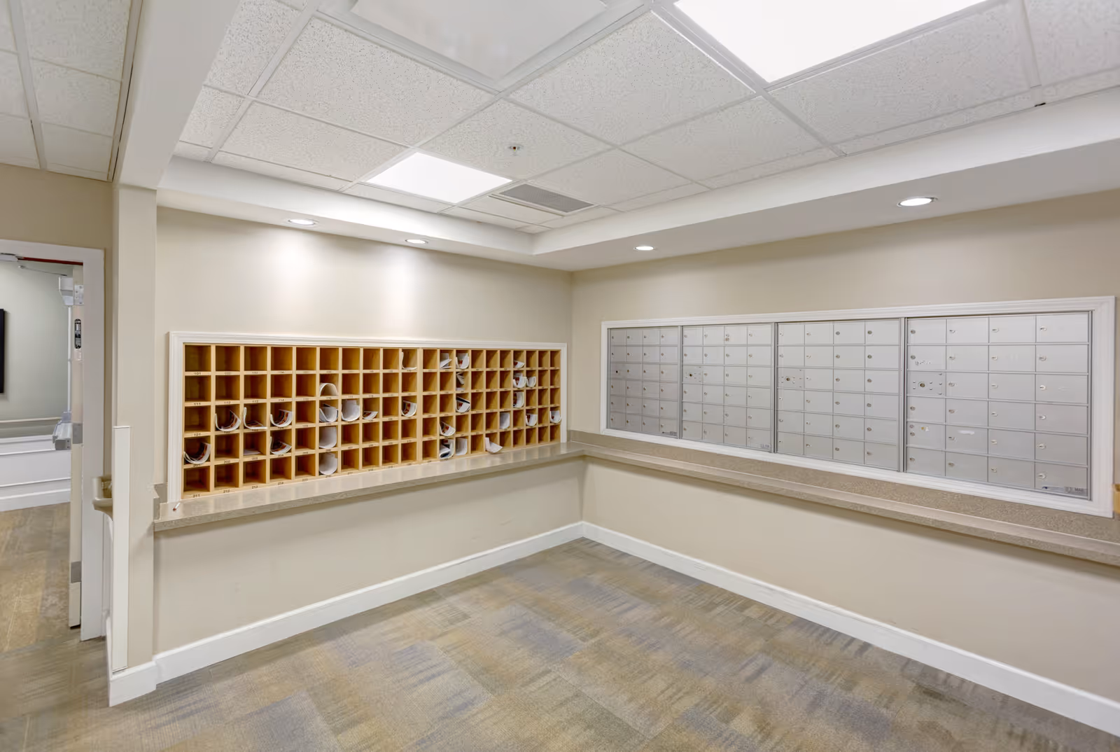 Interior view of a mailroom area with multiple small mailboxes on the right wall and a wooden cubbyhole organizer on the left wall, some containing rolled papers. The room has beige walls, a carpeted floor, and a drop ceiling with recessed lighting.