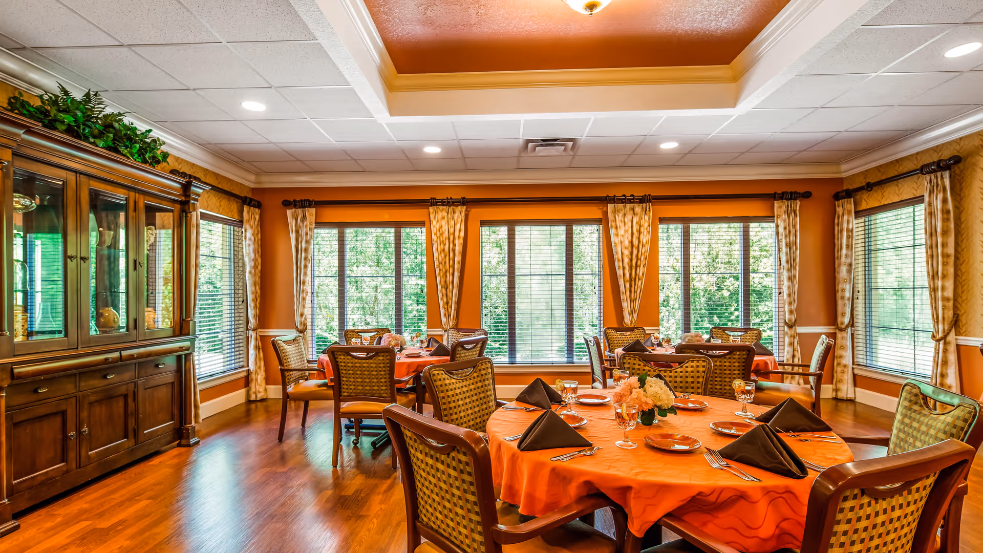 A bright dining room with large windows covered by curtains, round tables with orange tablecloths, neatly arranged place settings with black napkins, and wooden chairs. A wooden cabinet with glass doors is on the left side, and the room has warm orange walls and a white ceiling with recessed lighting.